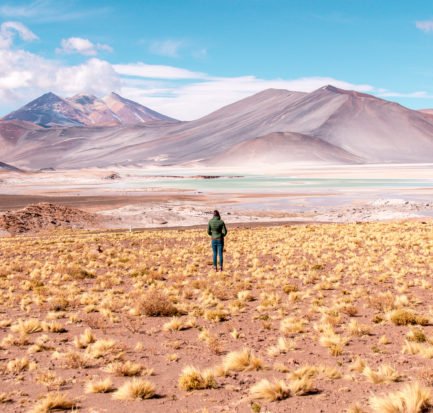 a woman in a green jacket standing in front of the purple mountains and blue lake in Atacama which is one of the most beautiful places in South America