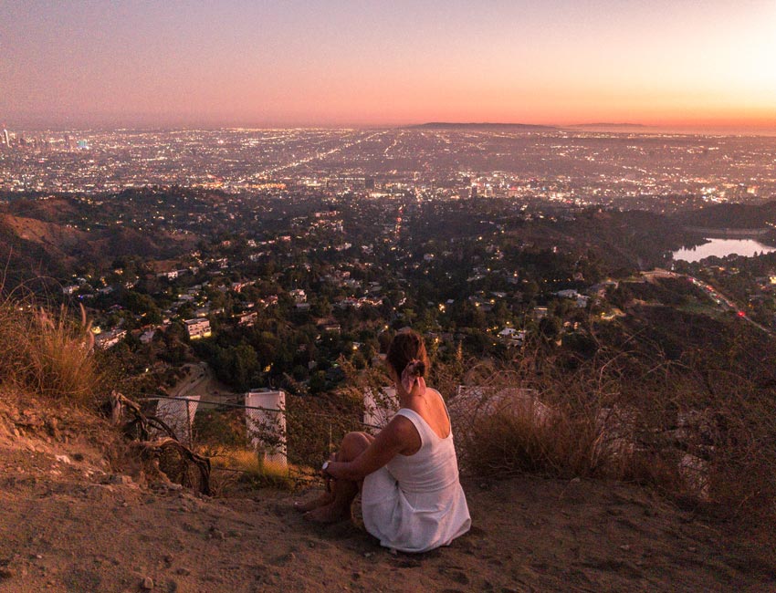 a girl in a white dress sitting on the ground behind the Hollywood sign during the sunset hour