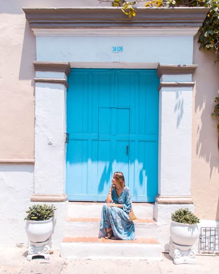 a woman in a blue dress sitting on the staircase in front of the blue door and two flower pots