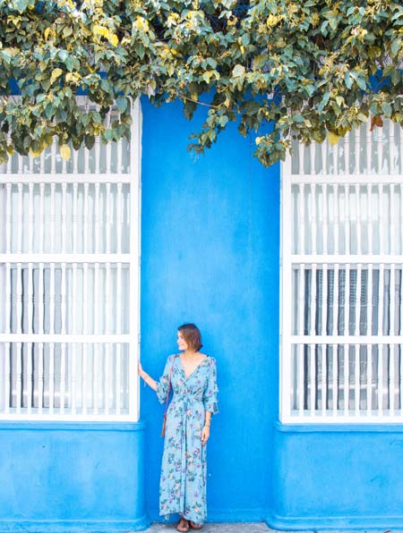 a woman in a blue dress standing in front of the blue wall , white windows and green plants