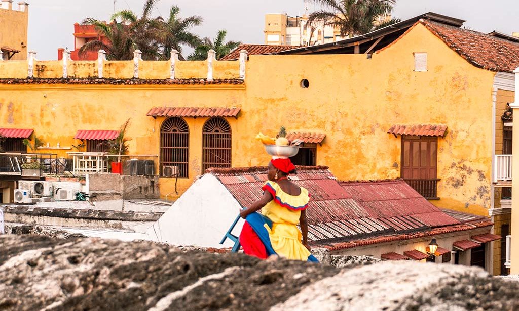 dreamy bucket list place called Cartagena, woman in a yellow dress walking in front of the orange building